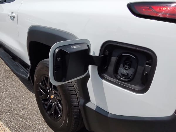 View of the left rear corner of a white EV truck with the door covering charging ports open.