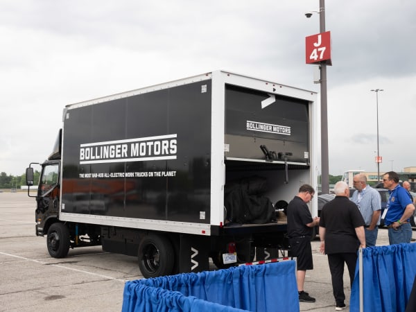 A Bollinger Motors electric truck is shown, with people chatting behind the truck.