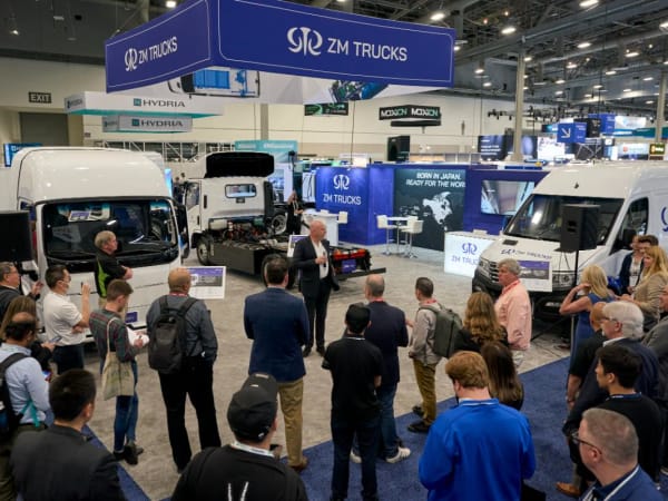 Media gather on show floor to listen to a press conference, trucks in background. 