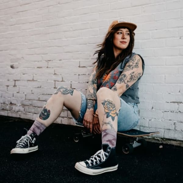 A young woman in jean shorts, wearing a baseball hat, leans against a brick wall. 