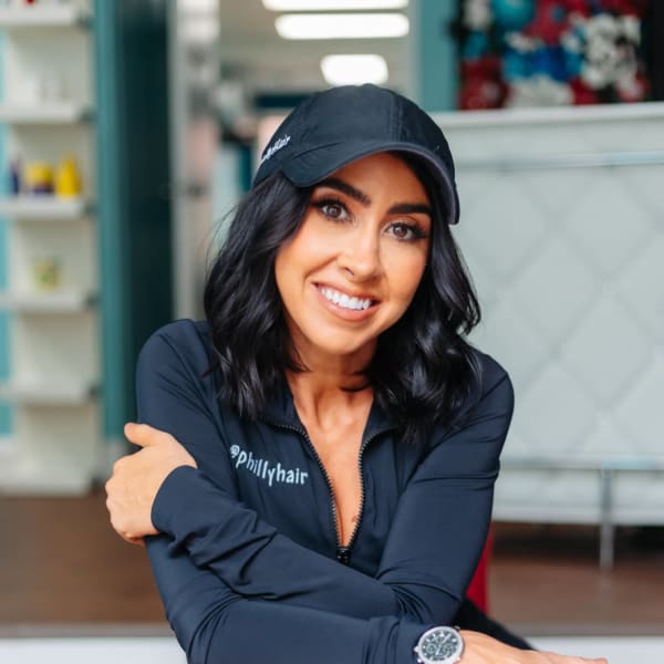 A woman wearing a black cap, leans on a red chair and smiles with folded arms.