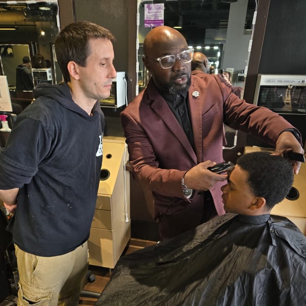 Man teaching a barbering technique to a student in a classroom setting.