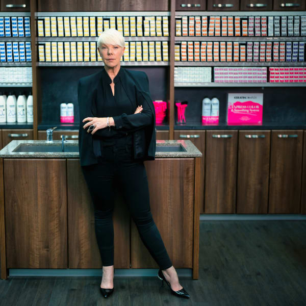 A woman in black and with short blonde hair, with arms crossed, standing in front of shelves of beauty products.