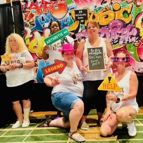 Five nail techs posing together in front of a branded selfie wall at the Nail Tech Event of the Smokies.