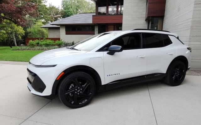 A white Chevrolet Equinox EV, small SUV, parked on the driveway outside a large, modern looking house.