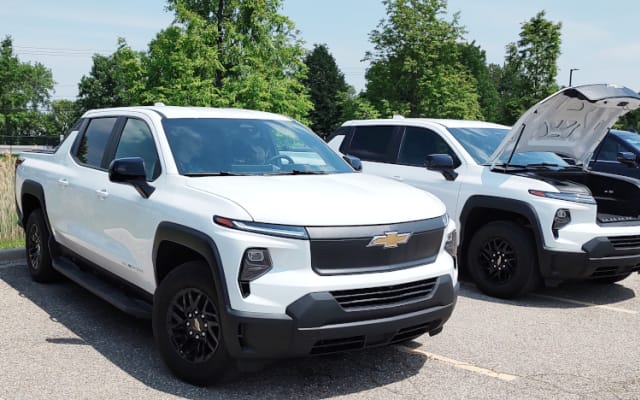 Two white EV pickups are parked, the one further from the camera has the front hood open.