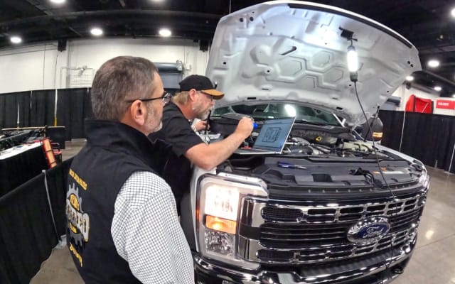 One man watching another work under the hood of a large pickup truck.