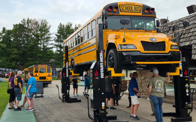 A new Blue Bird school bus is lifted on a heavy-duty Rotary vehicle hoist during an outdoor maintenance equipment demonstration as attendees gather nearby.