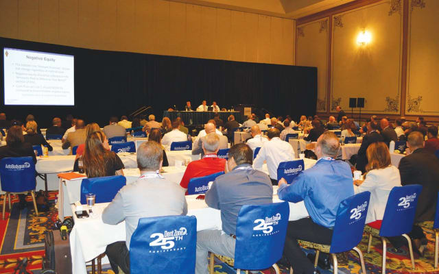 Audience seated at tables attending an automotive industry conference session, with a panel of speakers presenting on stage and a presentation slide displayed on a large screen.