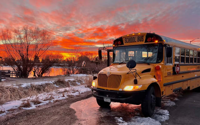 A yellow school bus is parked beside a snow-lined roadway at sunset, with vivid orange and pink clouds reflecting on a nearby pond. The bus's headlights and warning lights are illuminated against the colorful winter sky.