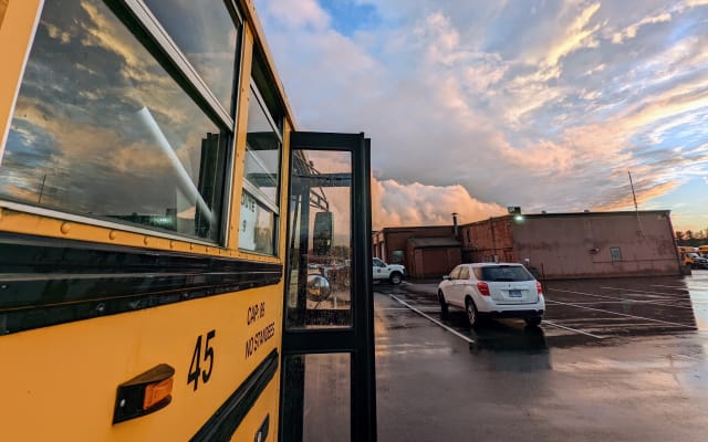 A yellow school bus with its service door open is parked after a rainstorm as dramatic clouds fill the evening sky. Reflections shimmer in the wet pavement, creating a scenic view of the bus yard at sunset.