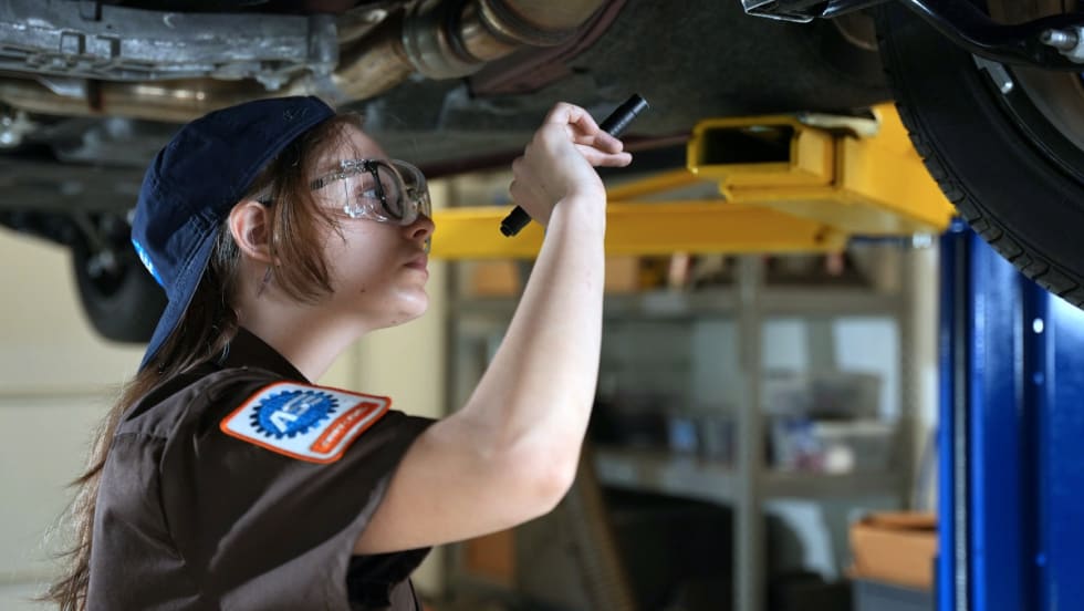 Female automotive mechanic uses a flashlight to check under a vehicle overhead on a lift.
