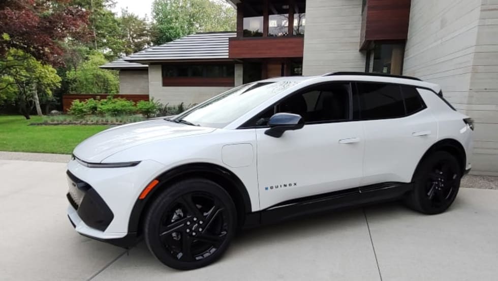 A white Chevrolet Equinox EV, small SUV, parked on the driveway outside a large, modern looking house.