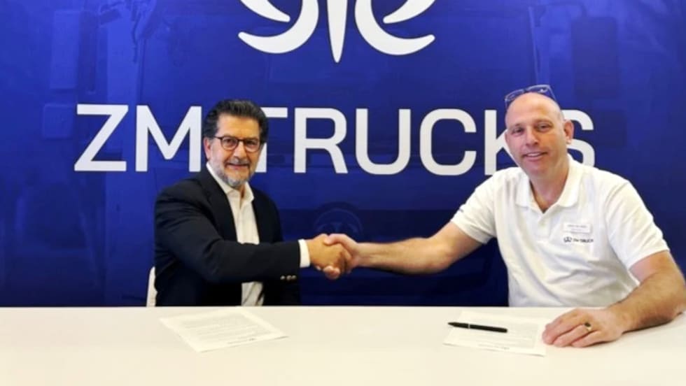 Two men seated at a table shake hands in front of a blue background with a logo for ZM Trucks.