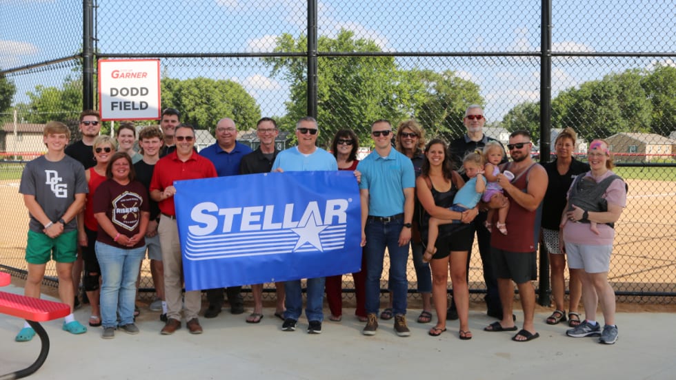 Group photo in front of backstop of a baseball field as people hold a banner that says Stellar.