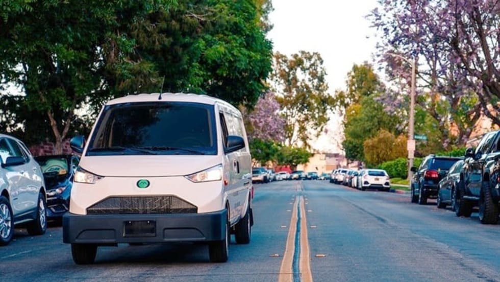 White EV van driving toward camera along shaded street.