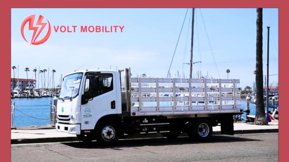 A Class 3 stake-body truck is parked in a parking lot in front of a marina, with the image also containing the logo for Volt Mobility.