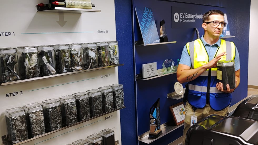 A man dressed in a safety vest is holding clear container of fine particles of black mass.