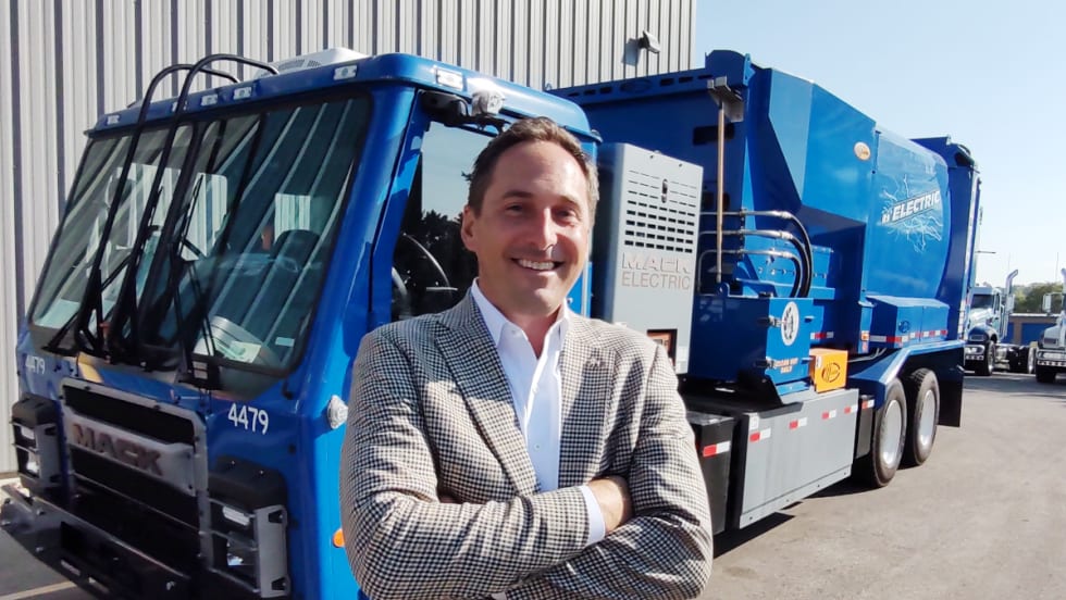 business attired man standing in front of a blue refuse truck