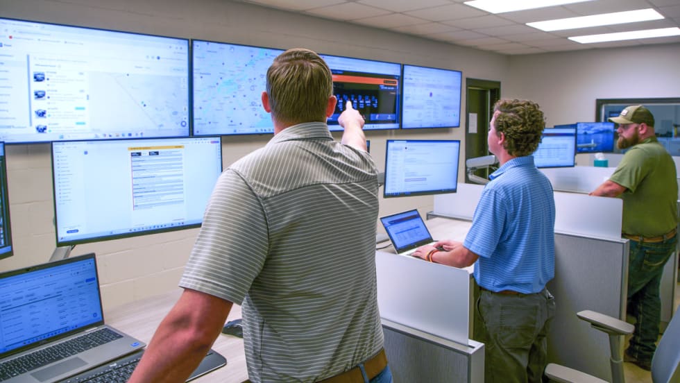 Group of men standing and monitoring computer screens.