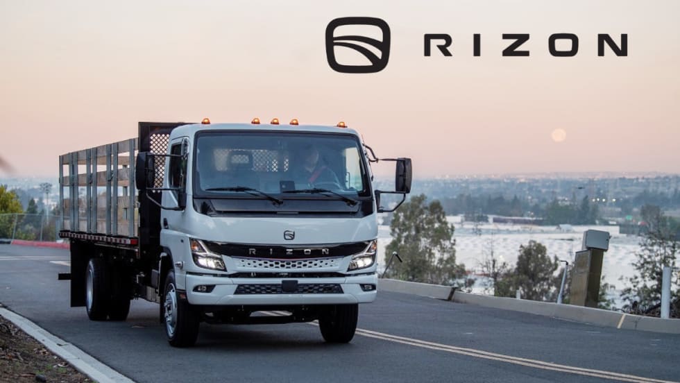 Stake body medium-duty electric truck driving on a road with sunset in background and Rizon logo upper right against sky.
