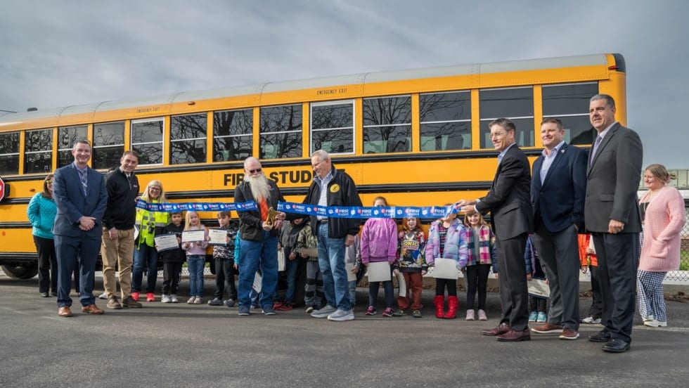 two people in safety vests walk in front of school buses