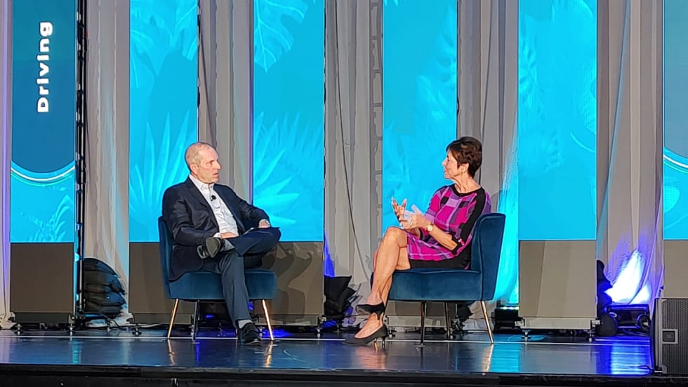 man and woman in business attire have a seated conversation on stage.
