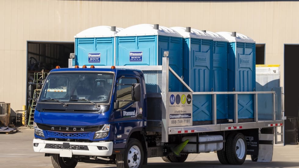 Battery electric truck with blue cab and flatbed hauling portable toilets.