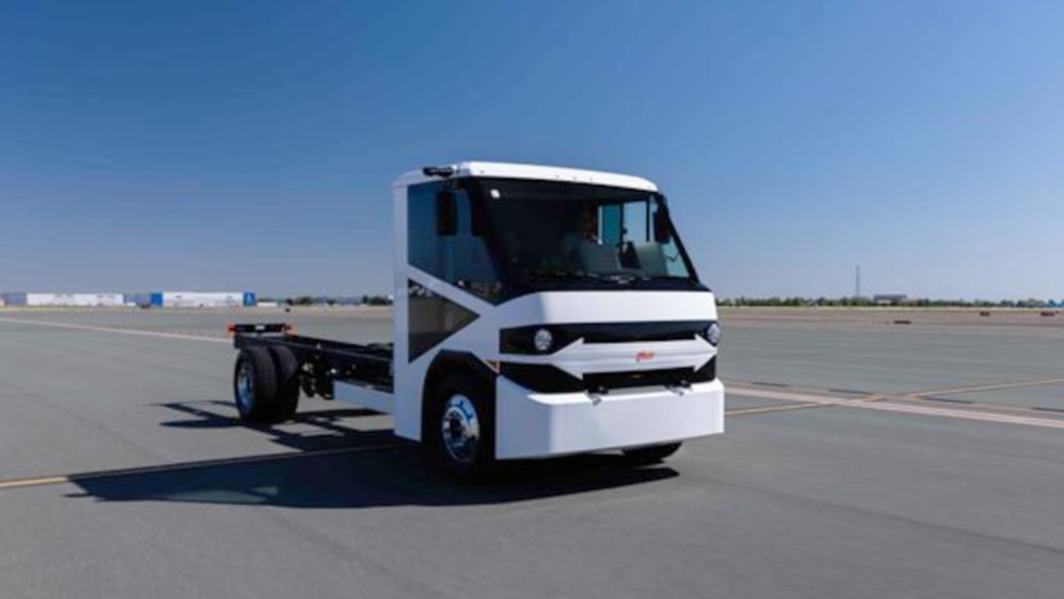 White battery-electric chassis cab parked in open paved area with blue sky.