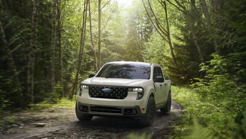A white 2025 Ford Maverick pickup driving on a dirt road in the woods.