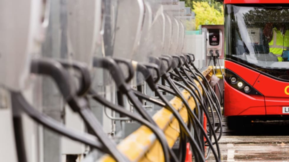 A row of electric bus charging stations with charging cables lined up, with a red double-decker bus plugged in and charging at the end of the row.