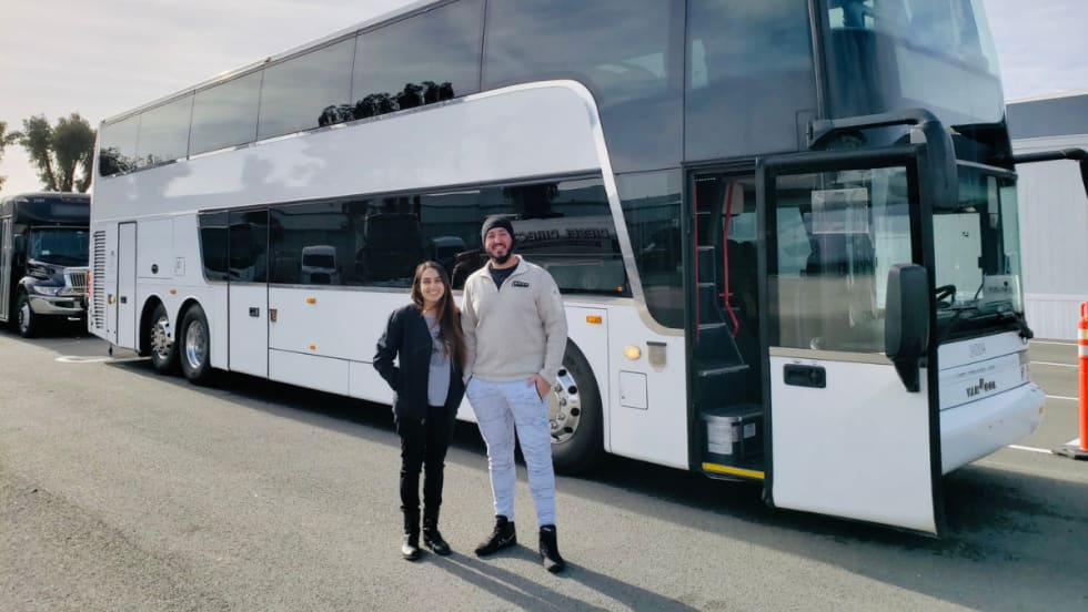 Two people standing in front of a white charter bus parked at a transportation facility.