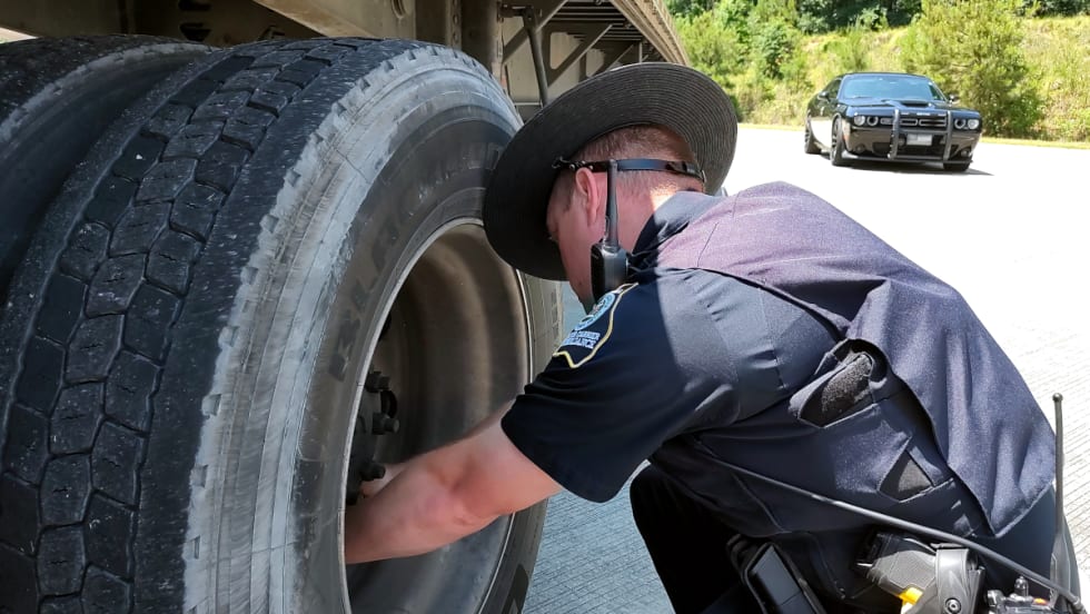 A state police officer inspects the wheels of a trailer with his patrol car parked in the background.