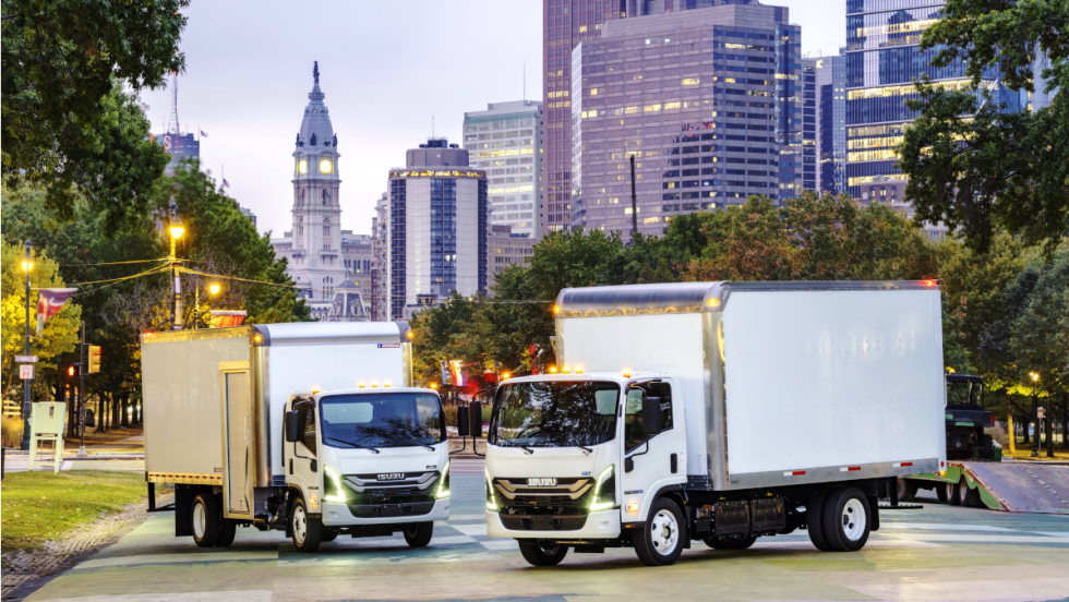 Two white Isuzu box trucks parked with a downtown skyline in the background.
