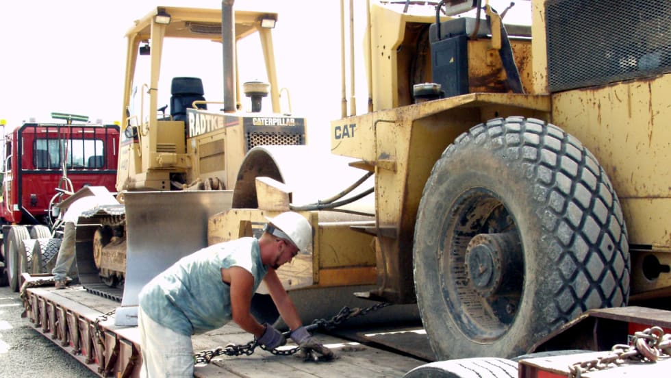 Construction worker tightens chain to secure a large piece of equipment on a trailer.