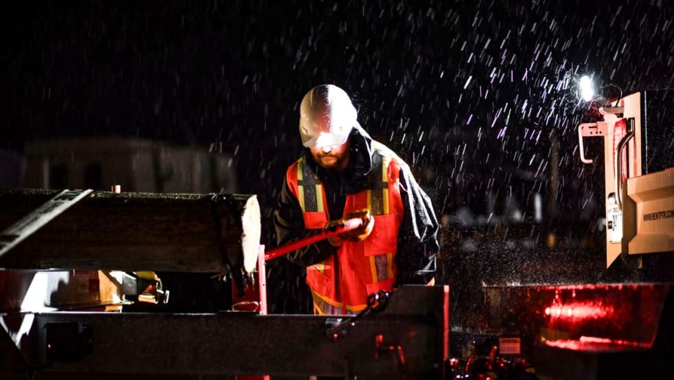 Worker wearing a high-visibility jacket and hard hat operates heavy equipment during a nighttime storm, with rain pouring down and debris secured on a trailer.