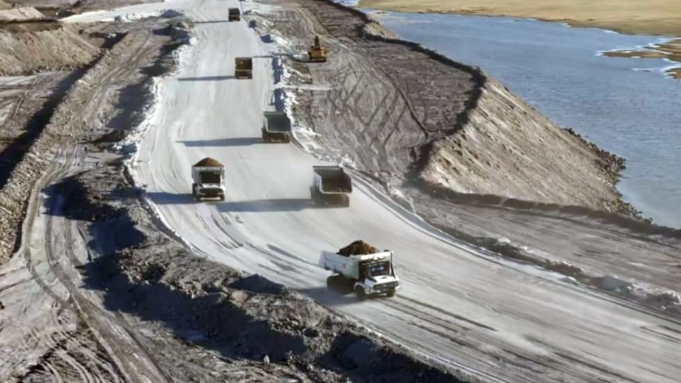 View of mining dump trucks driving on a curved road on top of a hill next to water.