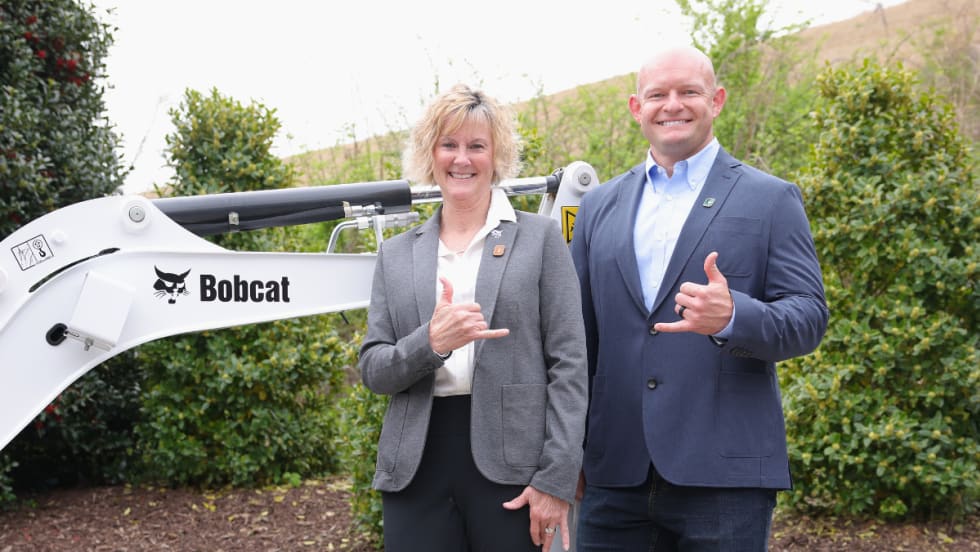A woman and a man in business attire pose in front of a Bobcat machine, smiling and making shaka hand gestures.