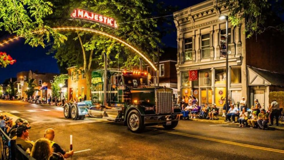 A classic Kenworth truck drives under a lit "Majestic" sign during the nighttime Kenworth Truck Parade in downtown Chillicothe, Ohio, as crowds line the street to watch the event.