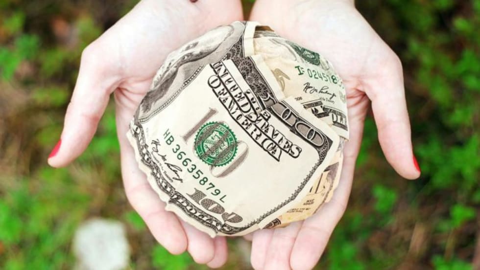 Hands holding a ball made of folded U.S. hundred-dollar bills, symbolizing financial assistance or giving.