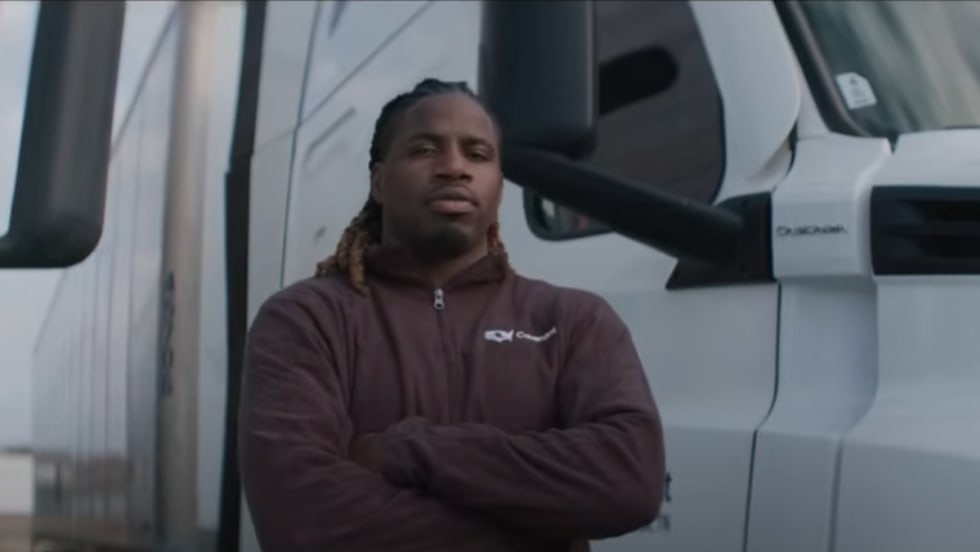 A truck driver stands with arms crossed in front of a white semi-truck, wearing a maroon jacket, featured in the ATA’s DRIVEN series highlighting professional drivers in the trucking industry.