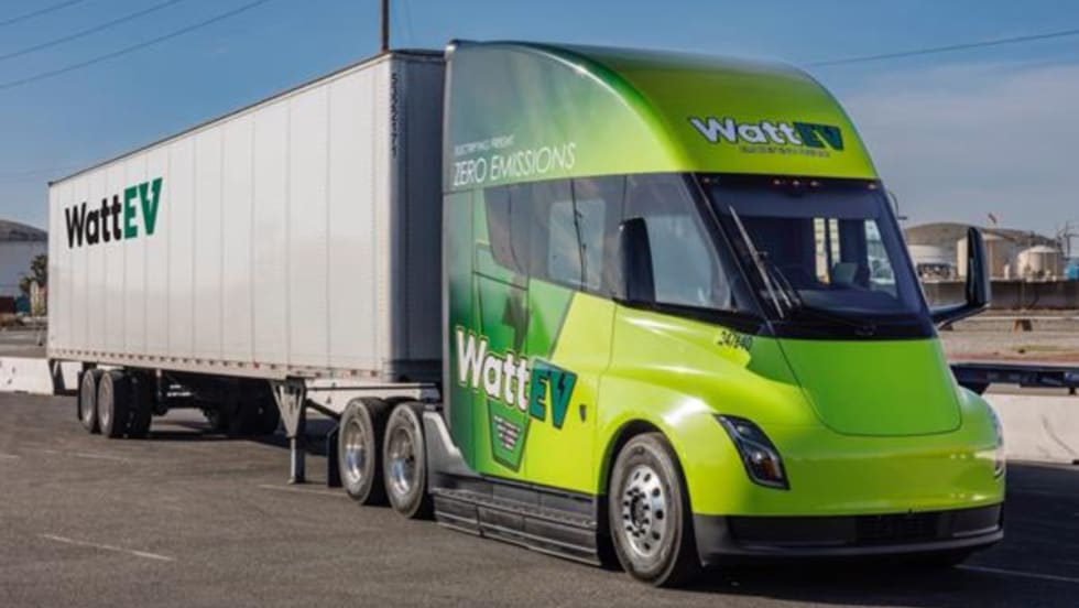 A WattEV electric semi-truck with a green cab and white trailer is parked in an industrial area under clear skies, showcasing the company's zero-emissions freight branding.