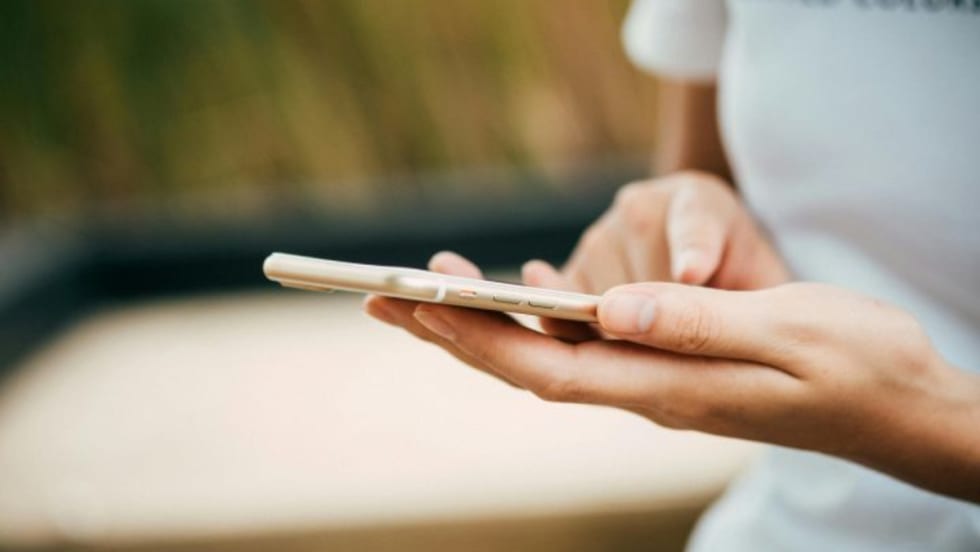 Close-up of a person using a smartphone, holding it with one hand and tapping the screen with the other, wearing a white shirt with blurred outdoor background.