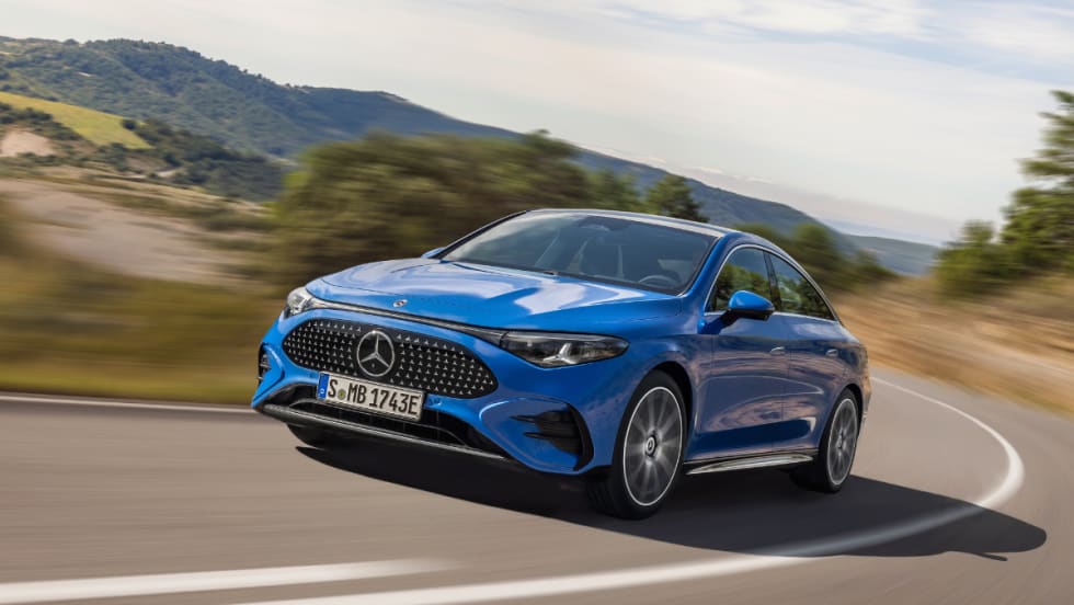 A blue Mercedes-Benz CLA drives on a curving rural road with hills and trees in the background, showcasing its aerodynamic exterior and daytime running lights.