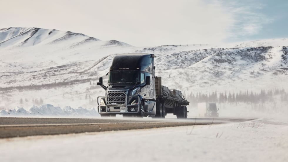 A Volvo VNL truck drives along a snow-covered highway in Fairbanks, Alaska, surrounded by mountains, as part of cold weather testing in extreme subzero temperatures.