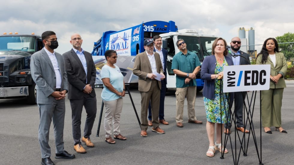 Group of officials standing around woman making announcement with two battery-electric trucks in the background.