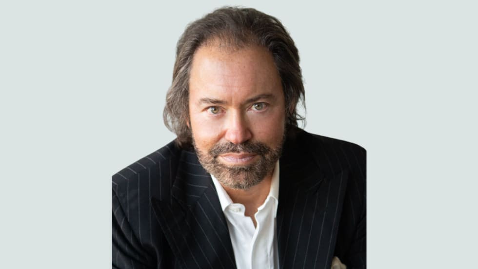 A man in a pinstripe suit jacket and white collared shirt looks directly at the camera with a neutral expression, seated against a light gray background.