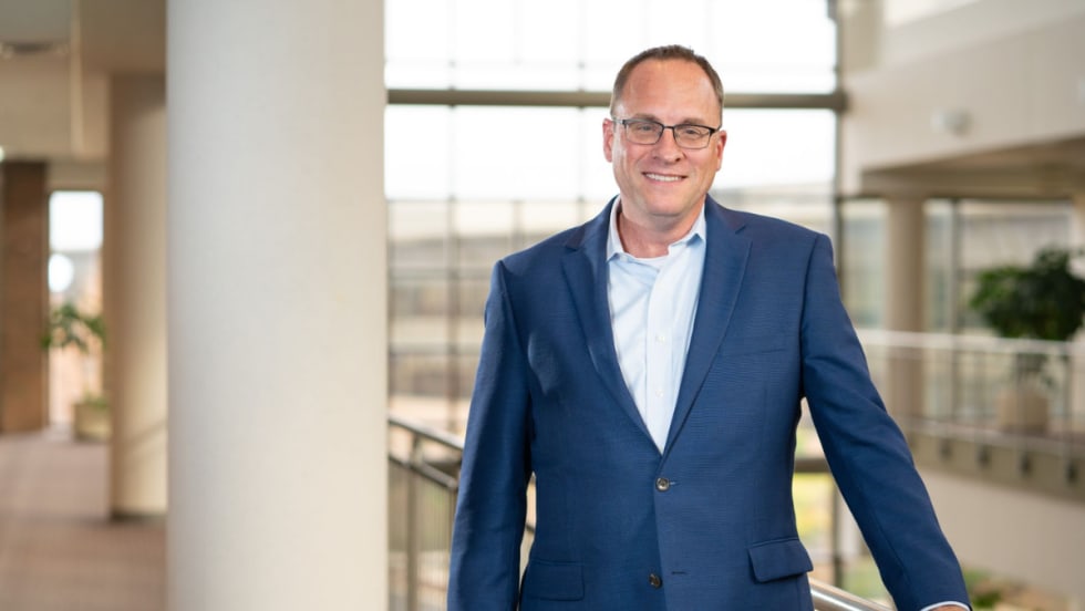 A man in a blue suit and glasses stands indoors near a railing, smiling, with large windows and natural light in the background.