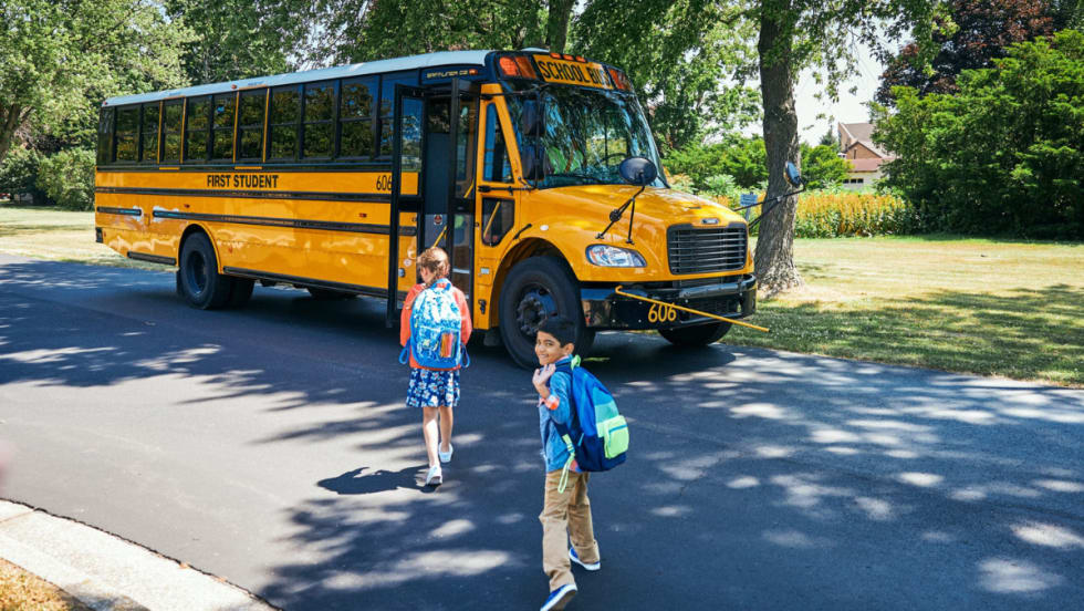 Two children with backpacks walk toward a First Student yellow school bus parked on a suburban street under the shade of trees.