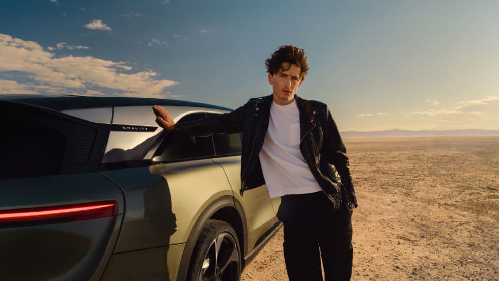 Actor Timothée Chalamet leans against a Lucid Gravity SUV in a desert setting, wearing a black leather jacket and white shirt, as part of Lucid’s new marketing campaign.